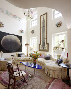 A bright living room with curved sofa, upholstered in light yellow floral fabric, eclectic art, tall windows, and colorful decorative accents.