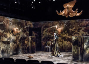 A theatrical stage set featuring forest mural walls, chandeliers, and dramatic lighting. A person stands in the center of the stage in a draped gown that matches the walls.