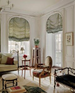 An elegant sitting room with arched windows, green window shades, carved moldings, and antique-style furnishings.