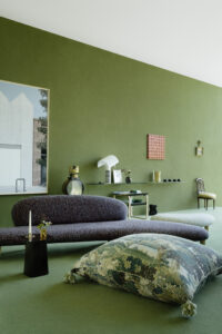 A contemporary green-walled room with a dark gray curved sofa, botanical floor cushions, sculptural lighting, and minimalist art accents.