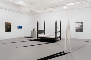 A minimalist gallery installation featuring a black four‑poster bed sculpture, stark white walls, and graphic floor elements.