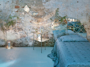 A softly styled bedroom featuring a weathered wall backdrop, blue bedding, mirrored accents, and delicate botanical details.