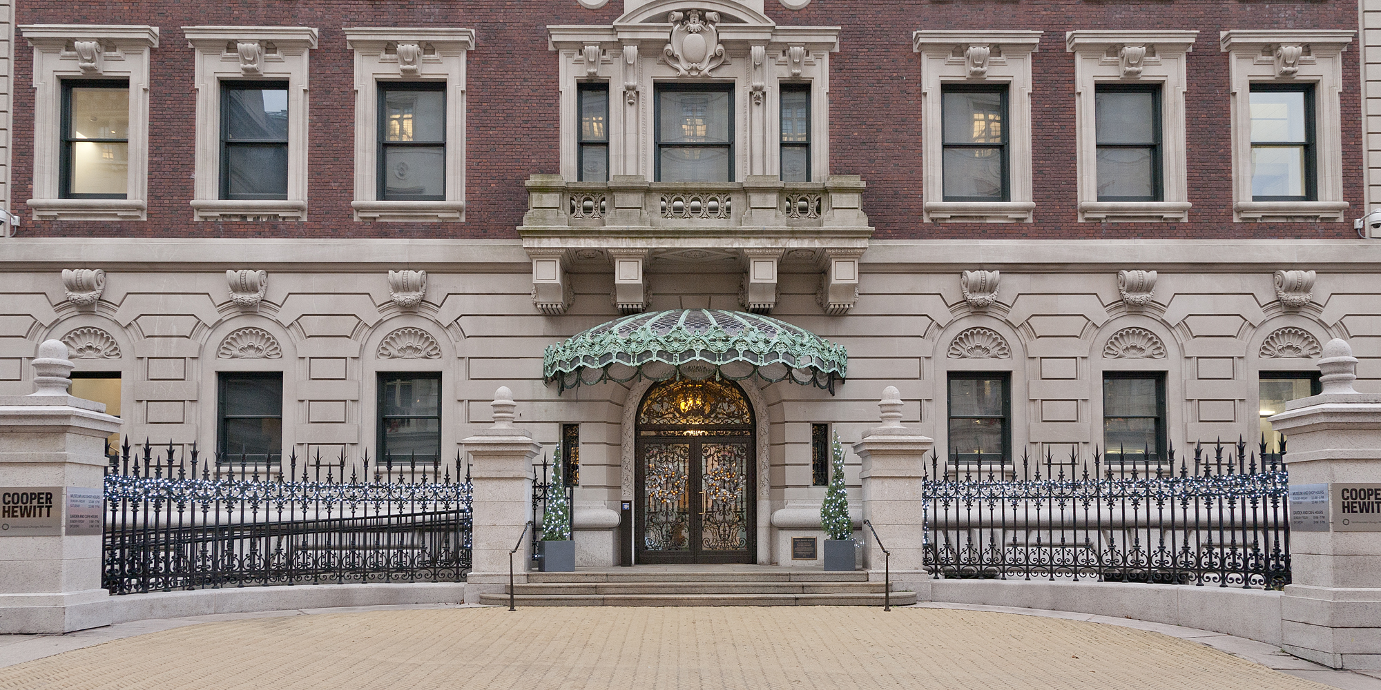 Grand entrance of the Cooper Hewitt, Smithsonian Design Museum: historic red-brick and light stone facade with ornate carvings, a large decorative doorway under a green-and-white striped awning, symmetrical tall windows with floral stonework, steps leading up flanked by small evergreen planters, wrought-iron fence, and a stone pillar engraved “COOPER HEWITT.”