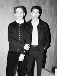Two individuals standing together against a wooden wall in a black‑and‑white photo.