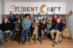 A group of ten people sit in front of a bookshelf. Text reads "Student Craft" in colorful letters on the back wall.