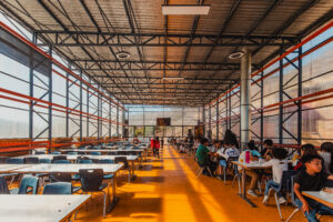 Large open dining hall with long tables, people eating together, and sunlight streaming inside.
