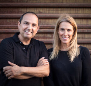 Two people in dark clothing standing together outdoors against a textured metal wall.
