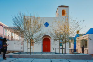 Modern white building with red arched doorway, trees in front, and people nearby outdoors.
