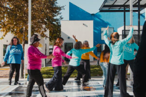 Group of people dancing outdoors on a tiled plaza beside colorful buildings and trees.