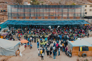 Large crowd gathered outdoors under a canopy in front of a multi‑story building.