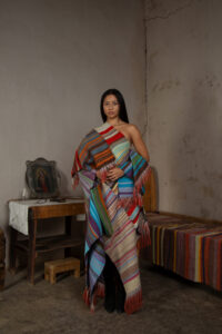 A model stands in a monastically-decorated concrete room dressed in a draped assemblage of striped cloth in many colors with fringe.
