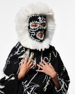 A person wearing an ornate black garment with white embroidered details and a dramatic circular headdress.