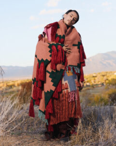 A model stands outside on a plain with a distant view of mountains dressed in a draped assemblage of geometric-patterned cloth in many colors with fringe.