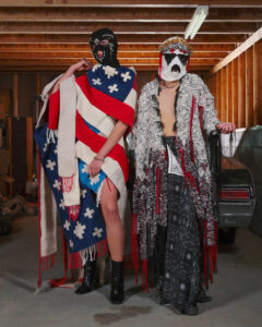 Two figures modeling avant-garde, handcrafted garments with bold textiles, masks, and dramatic fringe in a garage setting.