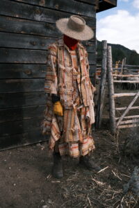 A person wearing layered patchwork garments, gloves, and a wide-brimmed hat beside a rustic wooden structure.