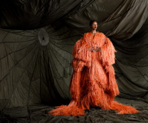 A person wearing layered, voluminous fringed garments in front of a black, draped background. The garments are a bright coral color.