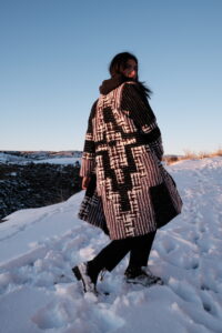A person wearing a black and white patchwork patterned woven coat stands in snow, overlooking a winter landscape.
