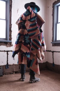 A person draped in a pink, purple, and blue geometric woven blanket and wide-brimmed hat inside a weathered room.