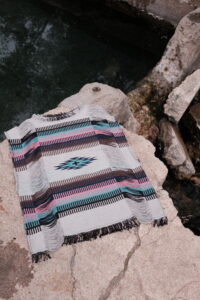 A woven geometric textile with fringed edges rests on stone beside dark, still water. The textile is white, black, brown, pink, and blue.