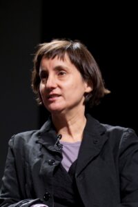 Person in dark layered clothing seated against a dark background, looking slightly to the side.