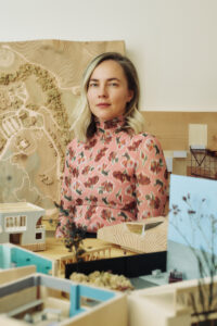 Person standing among architectural models and a carved landscape map in a studio setting.