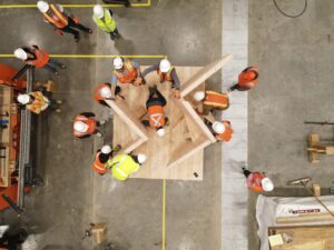 Birds eye view of construction workers assembling human-sized diagonal pieces of wood on top of larger flat rectangular piece.