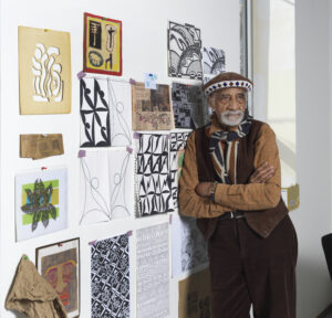 Person standing beside a wall covered in geometric drawings, patterns, and colorful mixed‑media artwork.