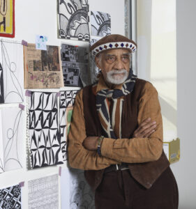 Person standing beside a wall covered in geometric drawings, patterns, and colorful mixed‑media artwork.