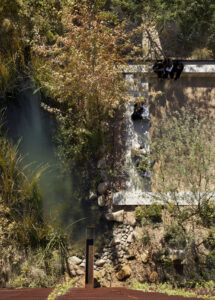 Birds-eye view of an outdoor courtyard with a seating deck, a natural pond, and surrounding desert flora.