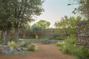 Outdoor courtyard with tables and chairs and decorated with desert fauna, walled in by a high fence made of stone pile and wire.