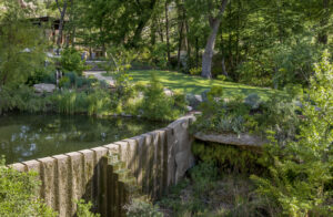 Lush green landcaped yard filled with towering trees and robust fauna that line a grass clearing next to a pond contained by a low moss covered stone fence with adjoining stone steps.