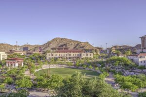 Sprawling campus centering a circular grass lawn with tan and terracotta school buildings, lush desert trees and fauna, and sidewalks radiating out from the center. Tan desert hills dotted with green surround the campus.