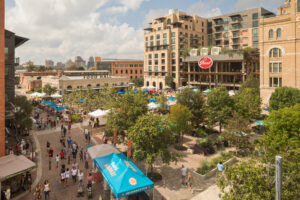 Outdoor plaza filled with trees and large crowds of people attending a famer's market called Pearl Market and a large indoor food hall, a towering building overlooking the plaza with a red sign near its roof reading "Pearl".