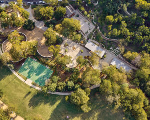 Birds-eye view of a sprawling outdooor greenspace that includes many activity centers, a large basketball court, and landscaped lush trees all bordered by an undulating white fence.