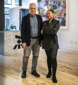 Two people standing in a bright art-filled room with wooden floors and colorful artwork.