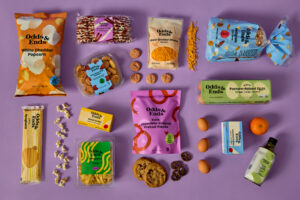 Various Odds & Ends food products in color-poppy, playful packaging arranged in an artful grouping against a lilac background.
