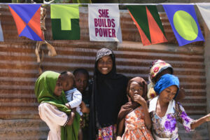 People gathered outdoors under bright flags and a banner reading “She Builds Power.”