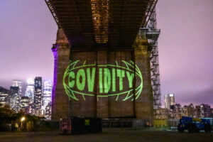 A large pillar supporting a bridge overpass displays the human-size projected words "Covidity" in neon green.