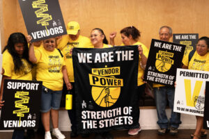 A line of people in yellow t-shirts that say "support vendors" joyfully displaying picket signs that read "we are the strength of the streets".