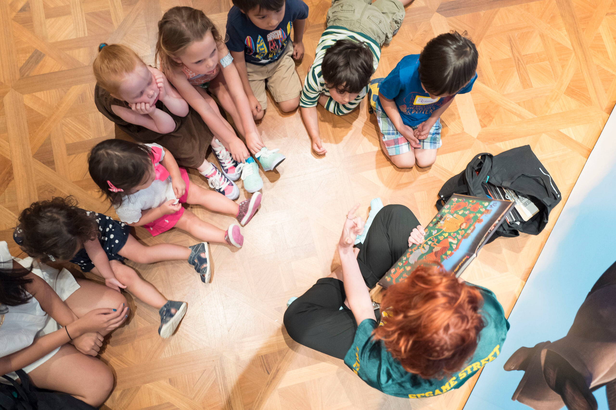 A circle of children sit on the gallery floor listening to an educator read a picture book.