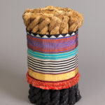 Cylindrical stool formed from an upright bundle of thick twisted rope sections dipped in black rubber at the base, banded around the middle with a vibrant, colorful patterned ribbon sleeve with orange fringe at the bottom.