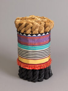 Cylindrical stool formed from an upright bundle of thick twisted rope sections dipped in black rubber at the base, banded around the middle with a vibrant, colorful patterned ribbon sleeve with orange fringe at the bottom.