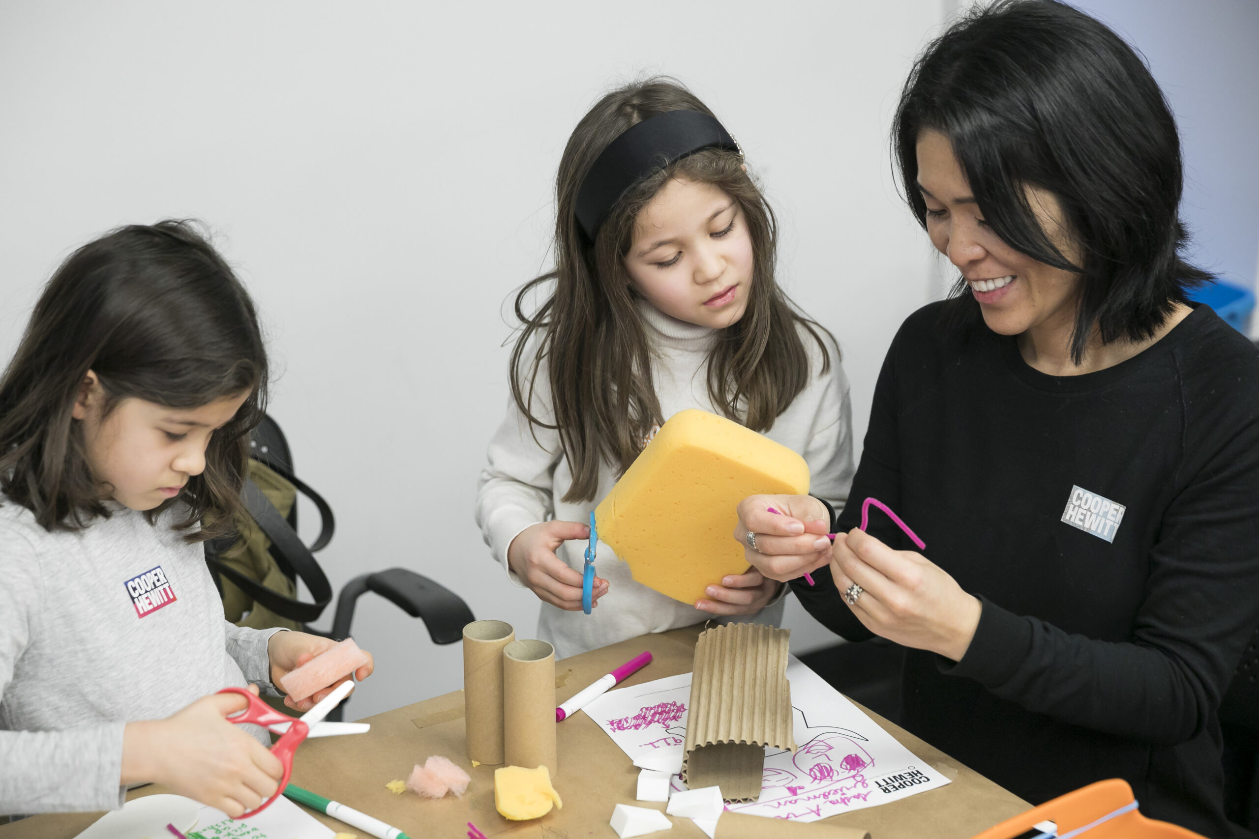 Two children sit at a table with their smiling mother and construct something out of materials