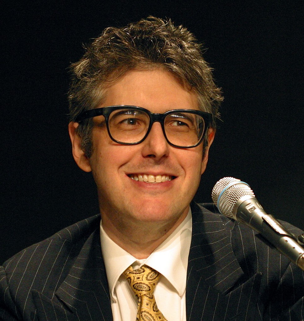 A photo of Ira Glass smiling and looking into the distance. There is a microphone in front of him.