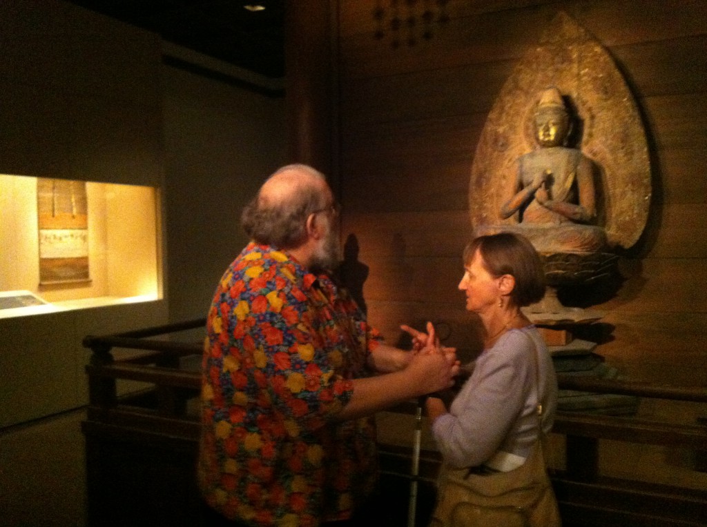 Dave and Linda are facing each other, standing a few feet in front of a Buddha statue. Dave is looking at the statue, and hoding Linta's arms. Linda is facing Dave and holding the pose.
