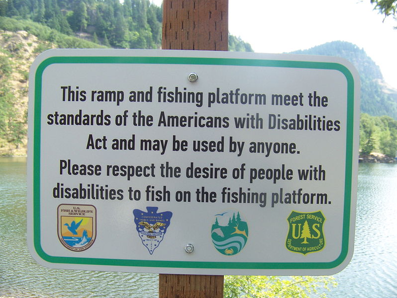 A sign in the foreground reads "This ramp and fishing platform meet the standards of the Americans with Disabilities Act and may be used by anyone. Please respect the desire of people with disabilities to fish on the fishing platform. In the background is a lake surrounded by trees.