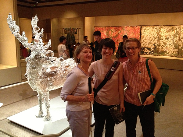 Linda, Rachel and Katie smiling inside a contemporary Asian art gallery at the Met museum. There is a very unusual sculpture in the background of a real deer, taxidermied and covered in glass orbs of variable sizes, as if it had been dunked in an oversized glass of club soda, and all the bubbles were sticking to its sides.