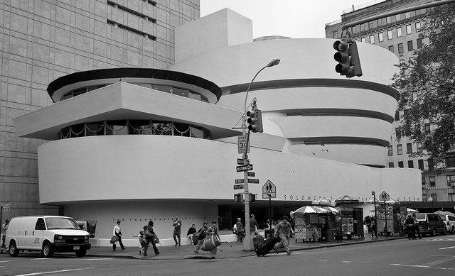 A black and white photo of the Guggenheim Museum in NYC. Traffic lights and pedestrians on the sidewalk are in the foreground. The museum's famous architecture looks like lots of big smooth white shapes stacked on each other: A big rectangle at the bottom, four big circles stacked on the right, and a second rotunda with windows on the left.