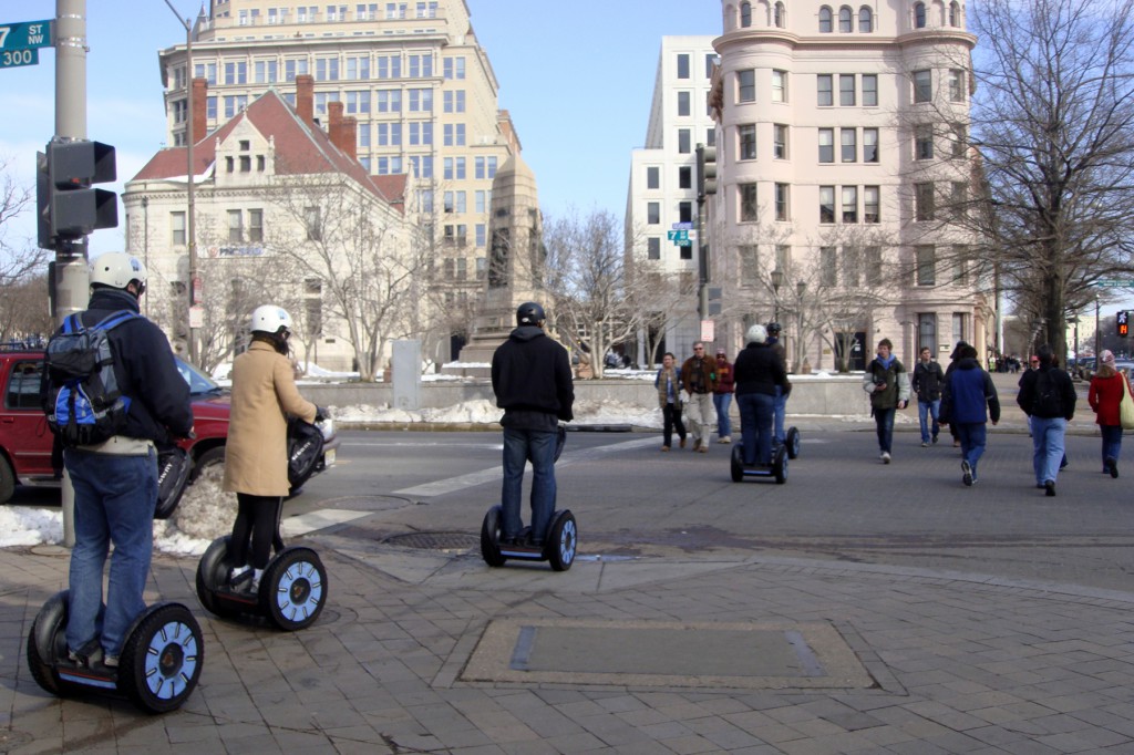 A group of five people riding motorized segway scooters riding single-file down the sidewalk curb cut and into the crosswalk. Washington DC in wintertime. They are wearing winter coats and helmets.