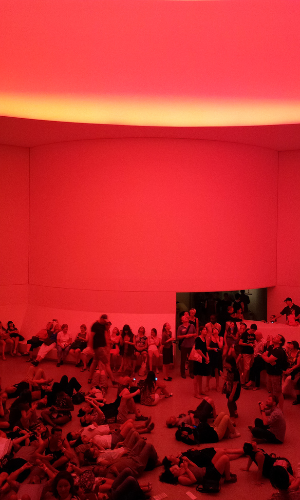 The Guggenheim's large round lobby, shown completely bathed in ruby-red light. The benches and floor area are crowded with people reclining, laying on the floor, and looking upwards at the light source.